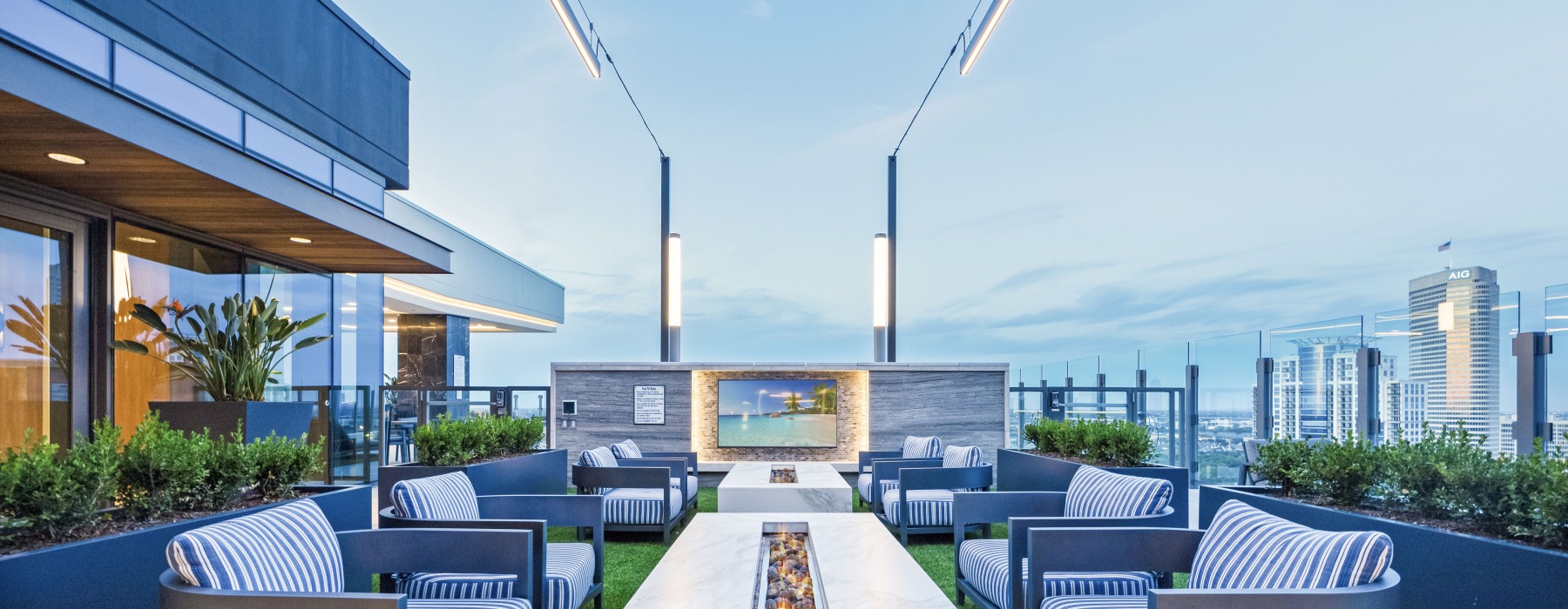 rooftop with low fire pit surrounded by cushioned arm chairs with a TV on a stone wall in the background